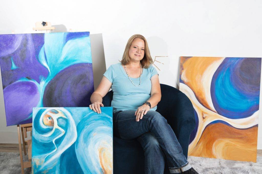 woman dressed in jeans and light blue t shirt sitting in plush chair with three abstract paintings around her smiling at the camera